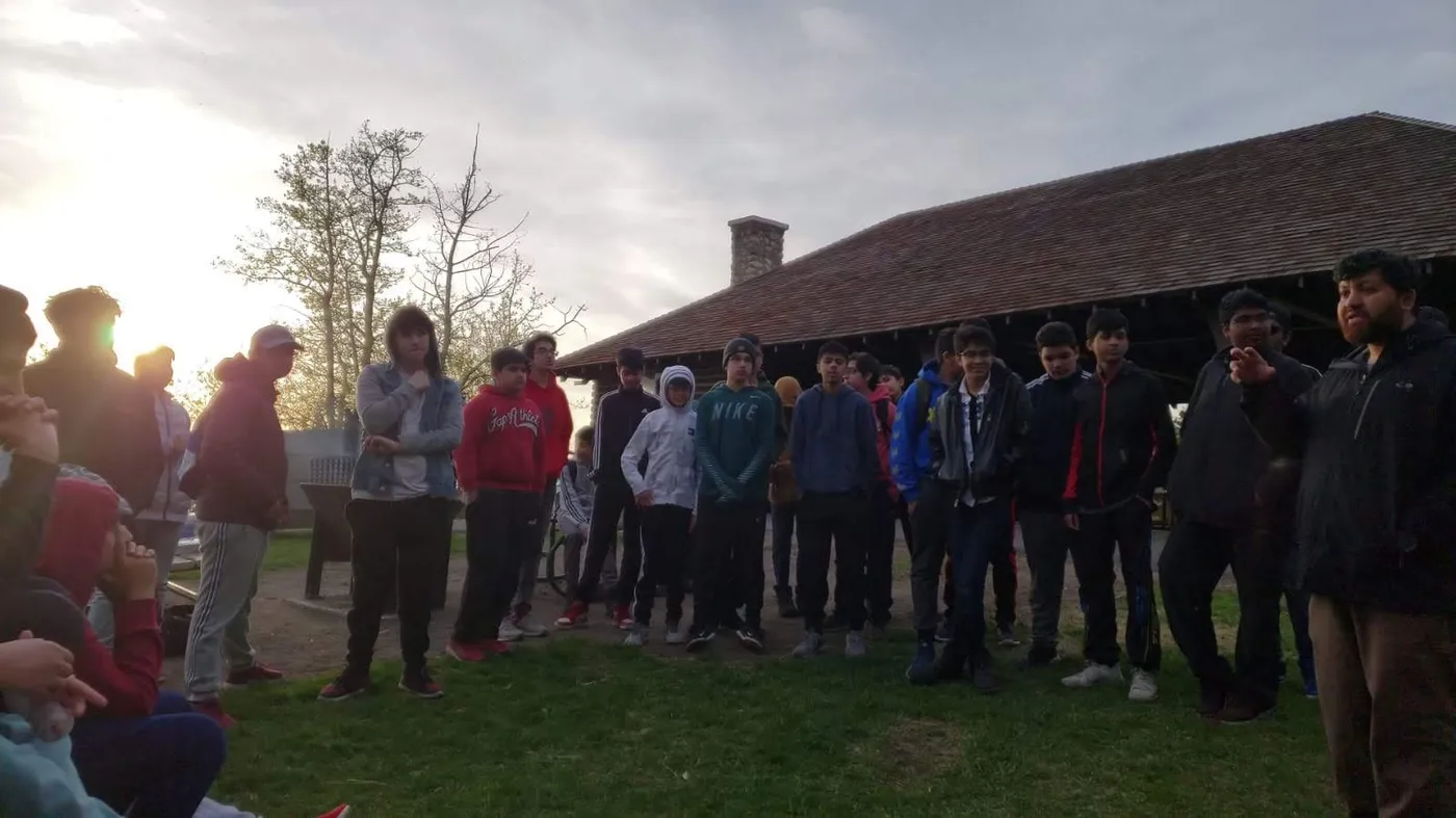 Dr. Qureshi speaking to a group of young people gathered on the grass outside a park shelter at dusk.