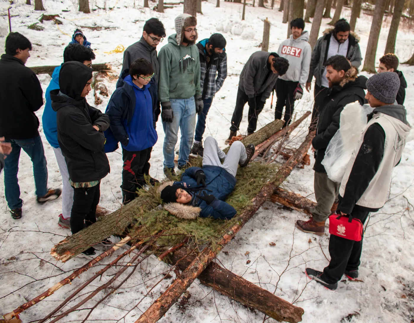 A winter first-aid exercise in the woods: students gathered around a casualty on an improvised stretcher of branches and boughs in the snow.